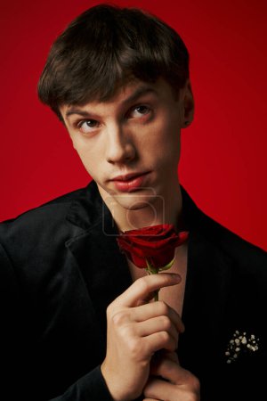 romantischer und verträumter junger Mann in Samtjacke mit Rose in der Hand auf rotem Hintergrund, Flirt