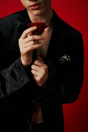 cropped photo of romantic young man in velvet jacket holding rose in hands on red background, flirt