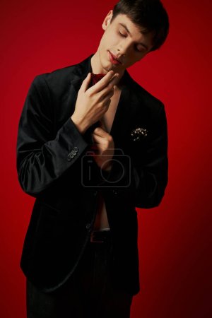 romantischer und verträumter junger Mann in Samtjacke mit Blick auf Rose in den Händen auf rotem Hintergrund