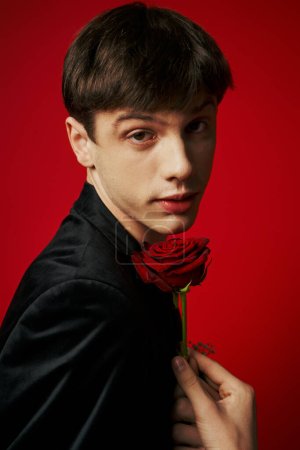 Porträt eines romantischen jungen Mannes in Samtjacke, der in die Kamera blickt und eine Rose auf rotem Hintergrund hält