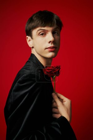 portrait of romantic guy in black attire looking at camera and holding rose on red background