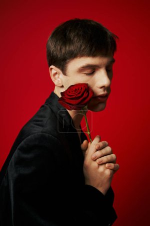 portrait of romantic guy with closed eyes holding rose near cheek on red background, flower