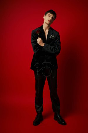 full length of romantic guy holding rose in hand and looking at camera on red background, flower