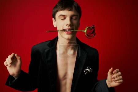 portrait of young man with closed eyes holding rose in teeth and smiling on red background, romance