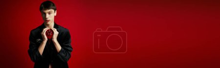 Téléchargez les photos : Homme tenant présent en forme de coeur et regardant la caméra sur fond rouge, bannière Saint-Valentin - en image libre de droit