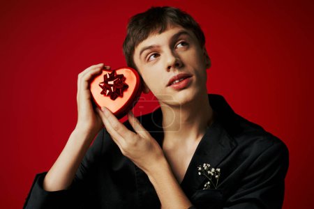 verträumter junger Mann im Samtblazer mit herzförmigem Geschenk auf rotem Hintergrund, Valentinstag