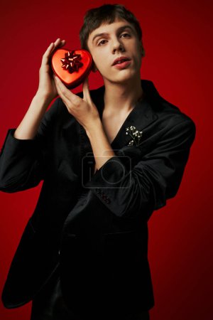 schöner junger Mann im Samtblazer mit herzförmigem Geschenk auf rotem Hintergrund, Valentinstag