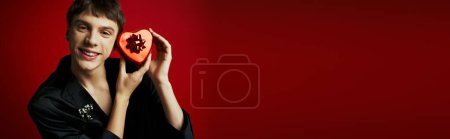 Téléchargez les photos : Homme gai en blazer de velours tenant présent en forme de coeur sur fond rouge, bannière Saint-Valentin - en image libre de droit