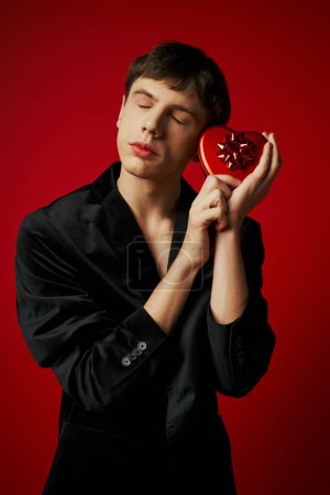 sentimentaler Mann im Samtblazer mit herzförmigem Geschenk auf rotem Hintergrund, Valentinstag