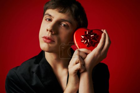 sentimental man in velvet blazer holding heart-shaped gift box on red background, Valentines day