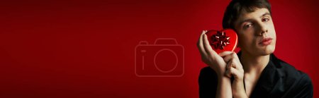 Téléchargez les photos : Charmant homme en blazer de velours tenant boîte cadeau en forme de coeur sur fond rouge, bannière Saint-Valentin - en image libre de droit