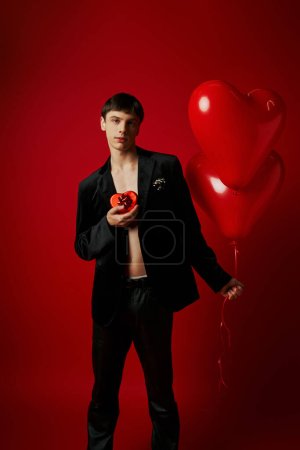 romantic young man holding heart-shaped gift box and balloons on red background, Valentines day