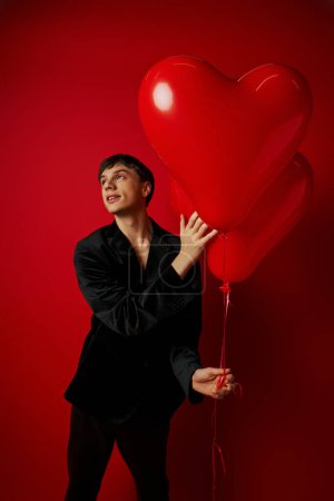 romantischer junger Mann im schwarzen Outfit mit herzförmigen Luftballons auf rotem Hintergrund, Valentinstag