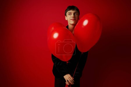 glücklicher junger Mann in schwarzer Kleidung mit herzförmigen Luftballons auf rotem Hintergrund, Valentinstag