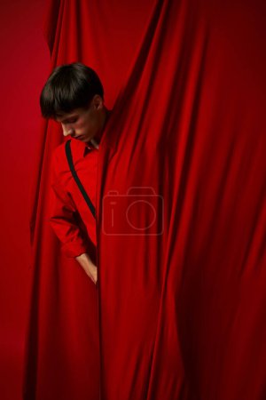 nachdenklicher junger Mann in pulsierendem Hemd mit Hosenträgern, der sich hinter rotem Vorhang versteckt, modischer Look
