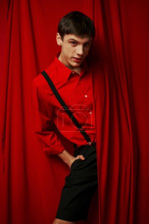 glücklicher junger Mann in lebendigem Hemd mit Hosenträgern, die sich hinter rotem Vorhang verstecken, modischer Look