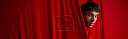 playful young man in vibrant shirt hiding behind red curtain while playing hide and seek, banner