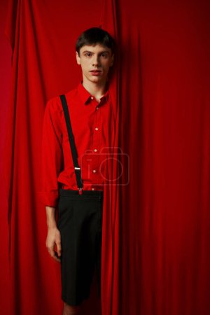 Photo for Young man in vibrant shirt and shorts with suspenders hiding behind red curtain, trendy look - Royalty Free Image