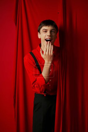 amazed young man in suspenders smiling and covering mouth near red vibrant curtain, merriment