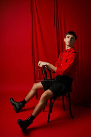 junger Mann in Hemd und schwarzer Shorts sitzt auf Stuhl in selbstbewusster Pose neben rotem Vorhang