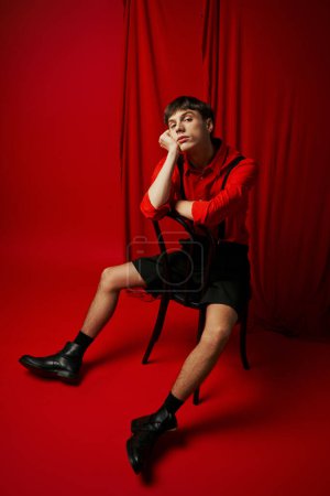 joven aburrido en camisa y pantalones cortos negros sentado en silla con pose relajada al lado de la cortina roja