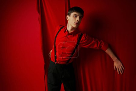 Foto de Hombre joven en camisa vibrante y pantalones cortos con tirantes de pie cerca de la cortina roja, aspecto de moda - Imagen libre de derechos