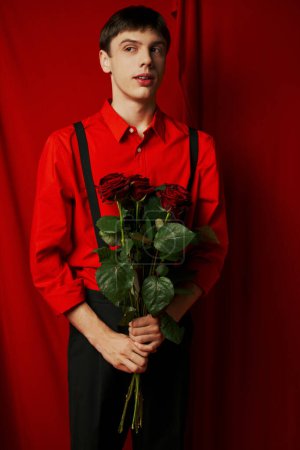 young man in vibrant shirt and shorts with suspenders holding bouquet of roses near red curtain