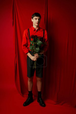 full length of young man in shorts with suspenders holding bouquet of red roses near bright curtain