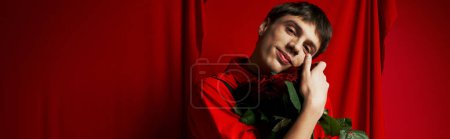 Téléchargez les photos : Heureux jeune homme en short avec bretelles embrassant bouquet de roses près rideau rouge, bannière - en image libre de droit
