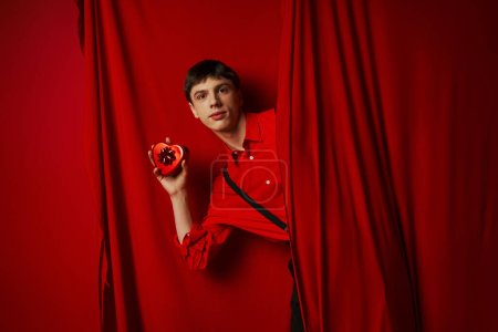 joven de camisa roja con tirantes sosteniendo caja de regalo en forma de corazón cerca de la cortina, 14 de febrero
