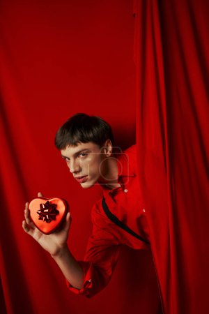 Téléchargez les photos : Jeune homme en chemise rouge avec des bretelles tenant coeur présent près du rideau, Saint Valentin - en image libre de droit