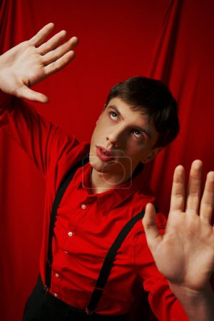 junger spannungsgeladener Mann im Hemd mit Hosenträgern posiert mit erhobenen Händen auf rotem Hintergrund, erschrocken