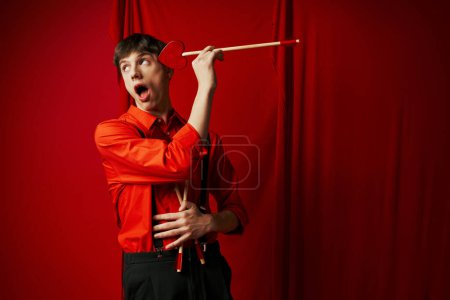 Schockierter junger Mann mit geöffnetem Mund und herzförmigem Pfeil auf rotem Hintergrund, 14. Februar
