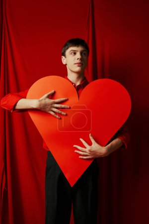 hombre joven alto en pantalones cortos negros abrazando gran recorte de corazón sobre fondo rojo, día de San Valentín