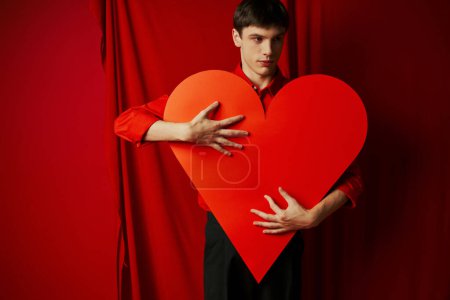 junger Mann in schwarzen Shorts umarmt großen herzförmigen Karton auf rotem Hintergrund, Valentinstag