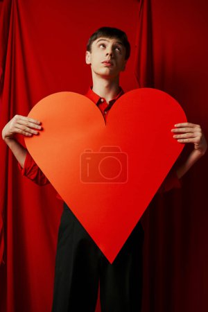 nachdenklicher junger Mann mit großem herzförmigen Karton auf rotem Hintergrund, Valentinstag-Konzept
