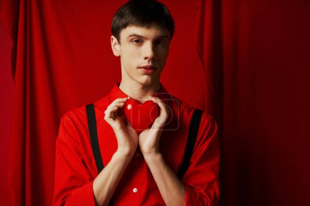 young man holding small heart in his hands and looking at camera on red background, 14 February