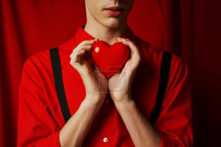 Téléchargez les photos : Vue recadrée du jeune homme tenant petit cœur dans ses mains sur fond rouge, concept Saint-Valentin - en image libre de droit