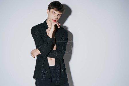 handsome young man in shiny black open shirt with hand near face posing on grey background