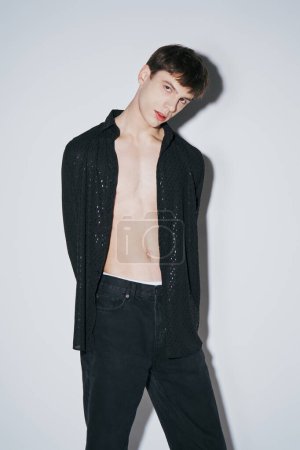 Foto de Joven guapo en camisa abierta negro brillante posando con las manos detrás de la espalda sobre fondo gris - Imagen libre de derechos