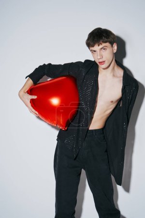 handsome young man in black shiny open shirt holding red balloon on grey background, 14 February
