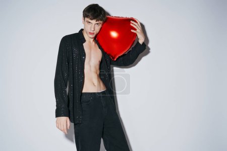 stylish young man in black shiny open shirt holding red balloon on grey background, 14 February