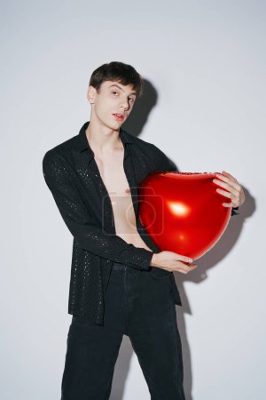 stylish young man in black shiny open shirt holding red balloon on grey background, Valentines day