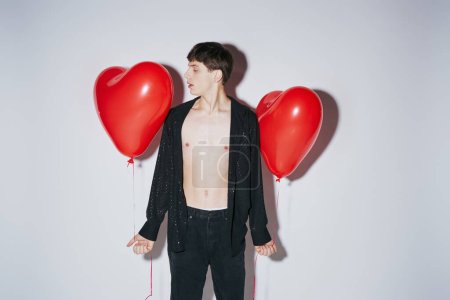 romantic young man in black shiny open shirt holding red balloons on grey background, Valentines day