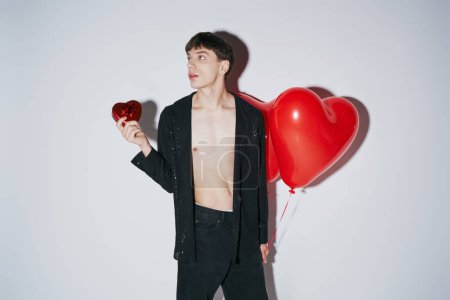 Mann im schwarzen Hemd mit rotem Ballon in Herzform und Geschenk auf grauem Hintergrund, Valentinstag