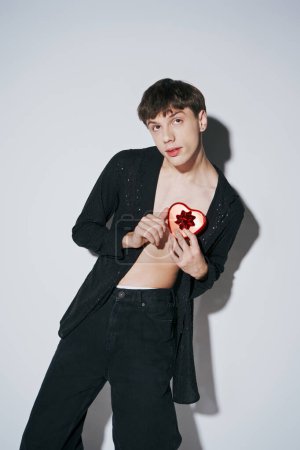 romantischer junger Mann im Hemd mit rotem herzförmigem Geschenk auf grauem Hintergrund, Valentinstag