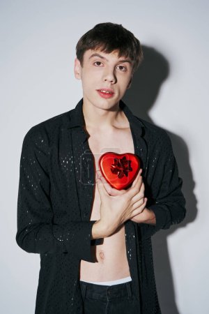 romantic young man in shirt holding red heart shaped present box on grey background, Valentines day