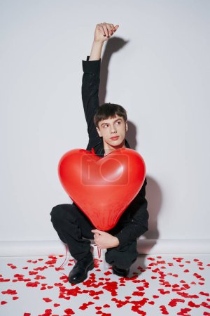 Foto de Joven levantando la mano y sosteniendo globo rojo cerca de confeti en forma de corazón sobre fondo gris - Imagen libre de derechos