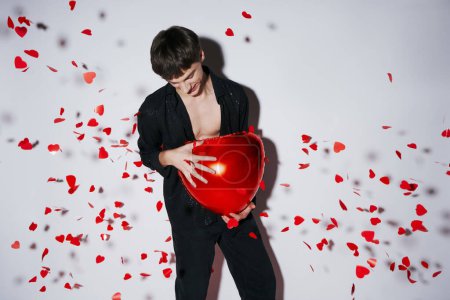 Foto de Joven en jeans y camisa sosteniendo globo rojo cerca de confeti en forma de corazón sobre fondo gris - Imagen libre de derechos