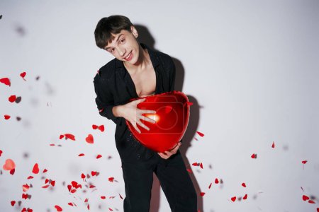 Foto de Hombre alegre en jeans y camisa sosteniendo globo rojo cerca de confeti en forma de corazón sobre fondo gris - Imagen libre de derechos